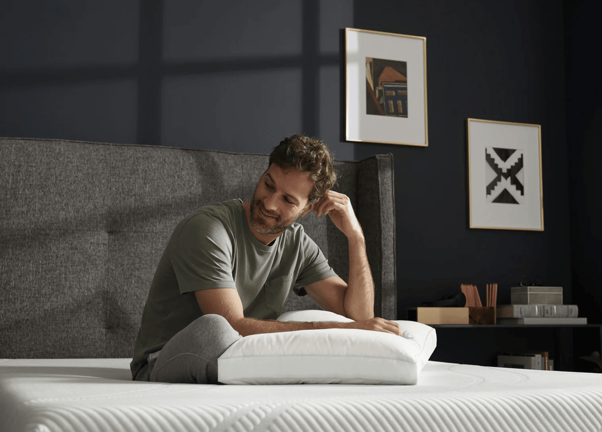 A man sits on a bed in a dark bedroom, holding a white pillow. The bed has a white quilted mattress, a gray upholstered headboard, and the wall behind it is dark navy blue with two framed art prints.