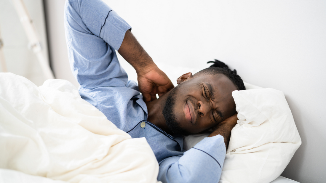 A man in a blue pajama shirt lies in bed, stretching and resting among white sheets and a light background.