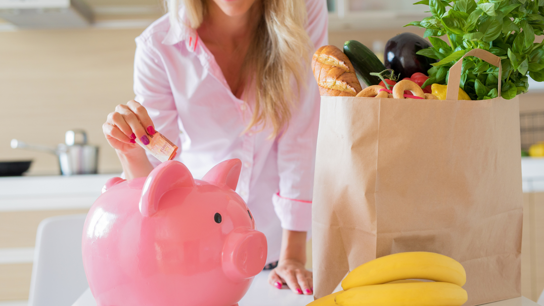 woman putting money into a piggy bank, next to a paper bag full of groceries