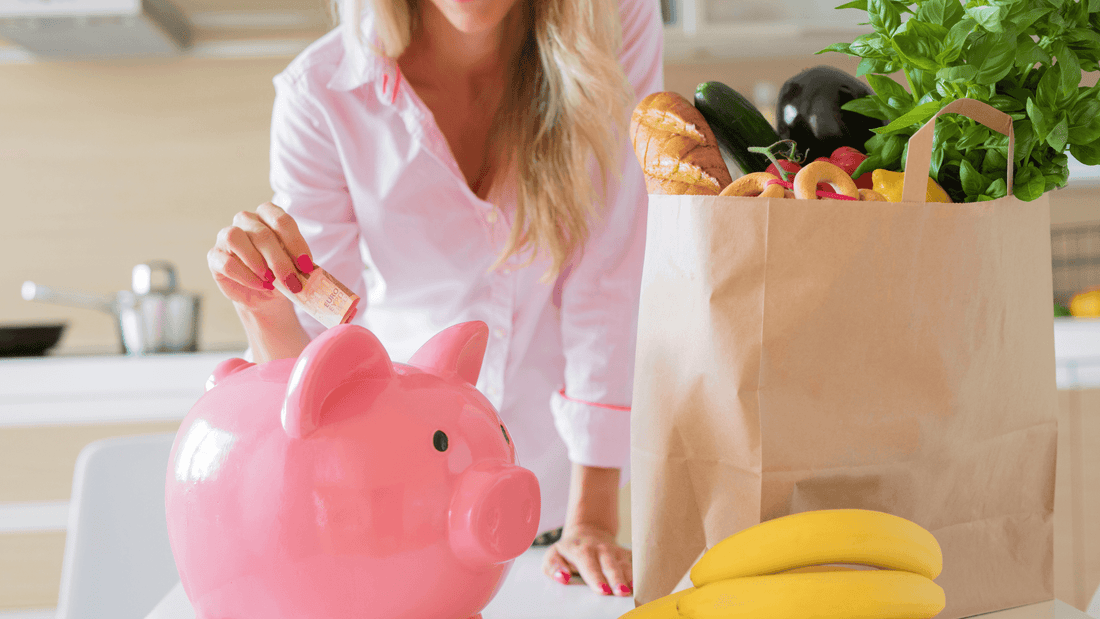 woman putting money into a piggy bank, next to a paper bag full of groceries