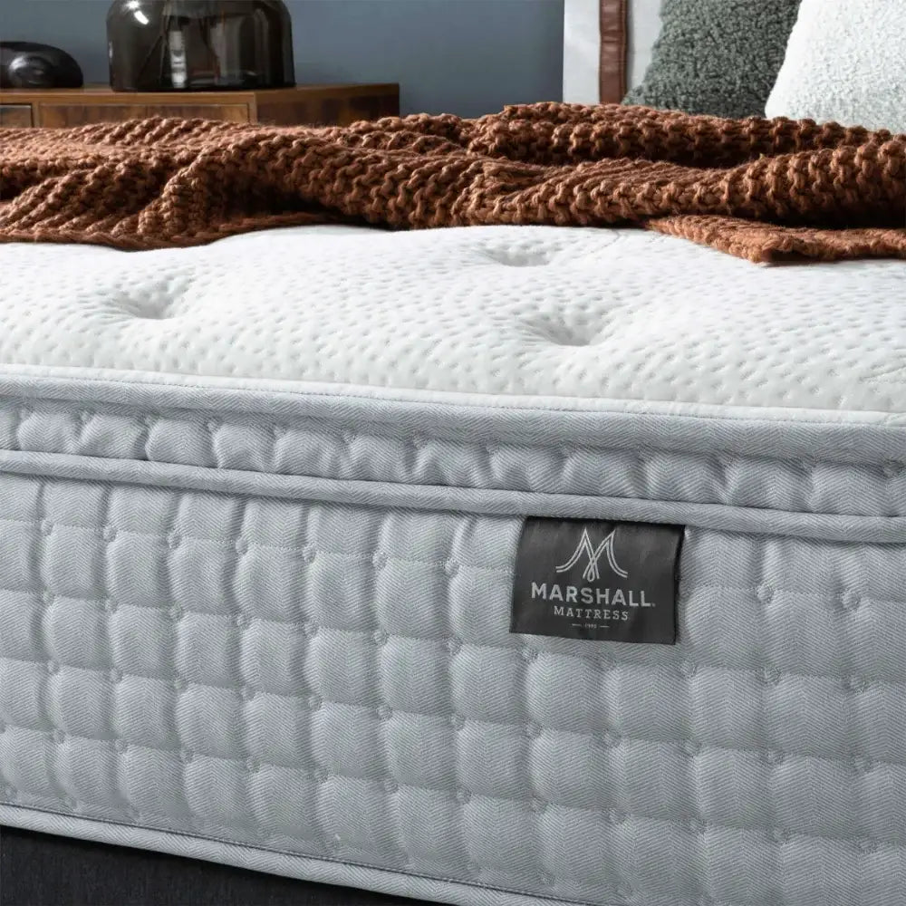 A close-up of Marshall Louise mattress with a tufted top surface, featuring a visible Marshall mattress label. A chunky brown knit blanket is draped across the upper portion of the bed, with a softly blurred bedroom background.