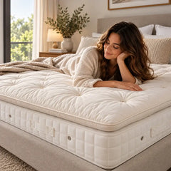 A woman in a cream knit robe lies on a sprout alpaca tufted mattress topper on a bed in a softly lit bedroom. Hypnos Mattress has a light beige side panel, with neutral bedding and a bedside lamp in the background.