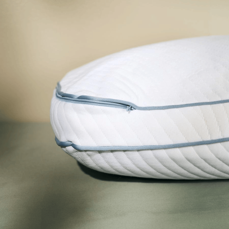 A close-up of a white quilted pillow with light blue piping and zipper detail, resting on a soft gray-green surface against a warm beige background.