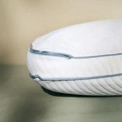 A close-up of a white quilted pillow with light blue piping and zipper detail, resting on a soft gray-green surface against a warm beige background.