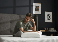A man sits on a bed in a dark bedroom, holding a white pillow. The bed has a white quilted mattress, a gray upholstered headboard, and the wall behind it is dark navy blue with two framed art prints.