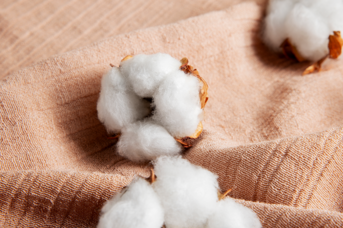 Close-up of fluffy cotton bolls on a soft, textured beige fabric. The scene conveys a sense of natural softness and warmth.