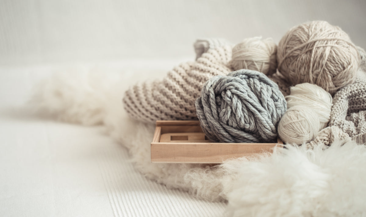 Cozy scene with skeins of beige and gray yarn in a wooden tray, surrounded by soft knitted fabric and a fluffy white rug, conveying warmth and relaxation.