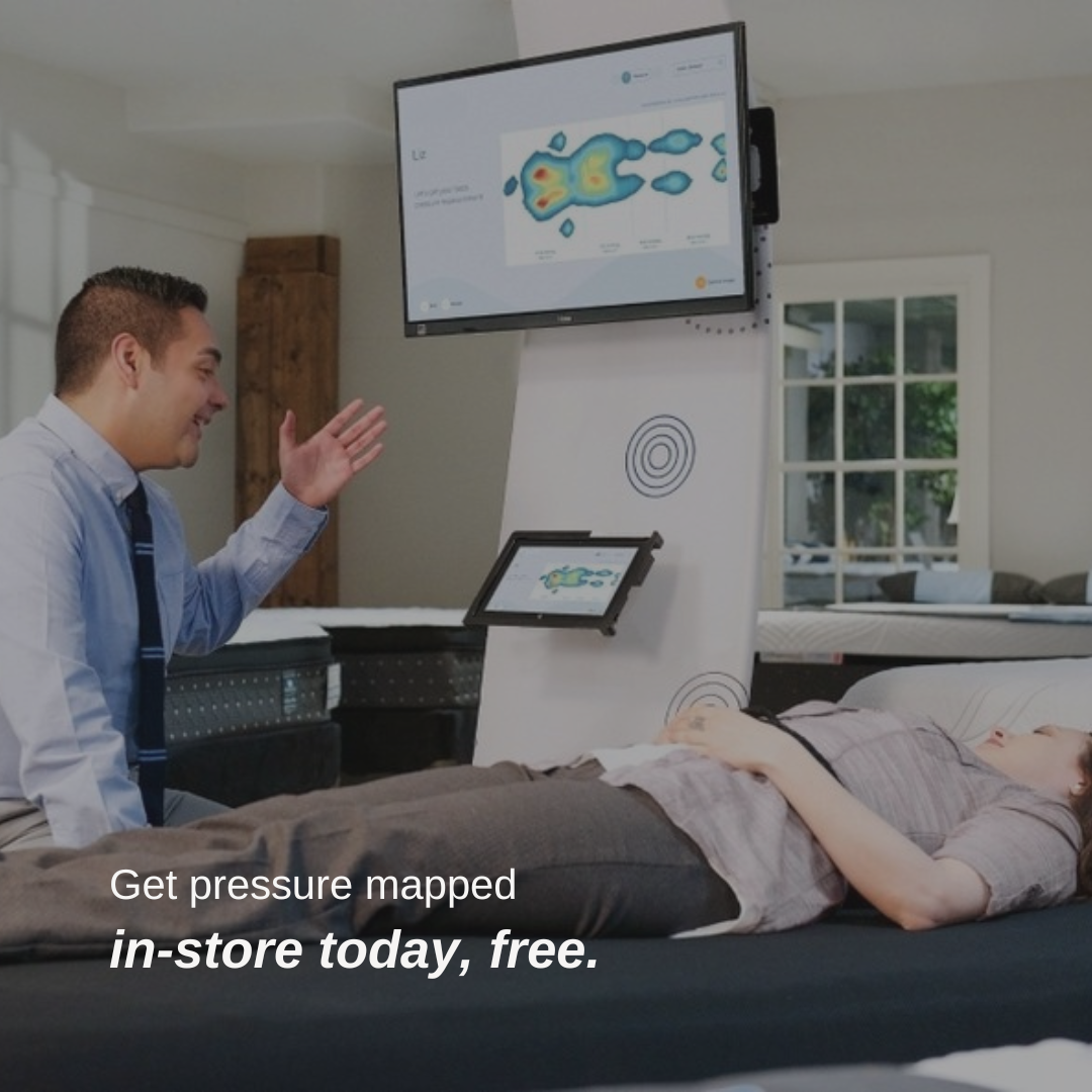 A store employee explains a mattress pressure mapping system to a customer laying down. A screen displays pressure areas. Text reads: "Get pressure mapped in-store today, free."-image