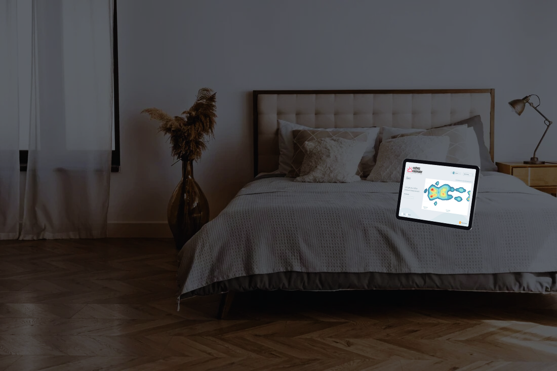 A cozy, minimalist bedroom with a tablet displaying a heat map on the bed. Soft lighting, pillows, a bedside lamp, and a vase with pampas grass enhance the calm ambiance.
