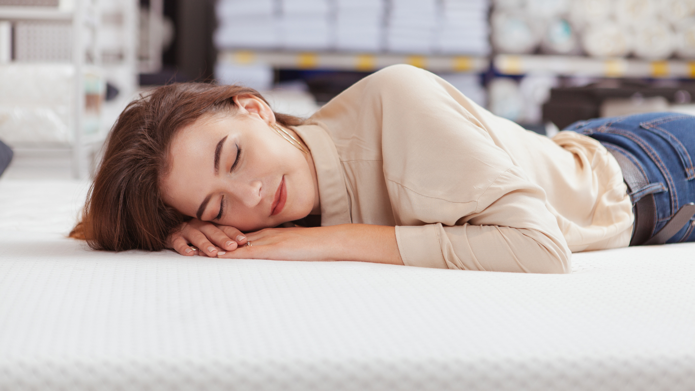 Woman lying on a white mattress with closed eyes and a gentle smile in a store setting.